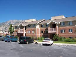 A two-story apartment building with red brick and tan siding, balconies, and a landscaped front yard sits in Wyview Park Provo. Several cars are parked in a lot, with mountains visible under a clear blue sky.