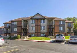 The Branbury Provo offers a two-story apartment building with brown and beige siding, outdoor staircases, and balconies. Several cars are parked in the lot under a clear blue sky.