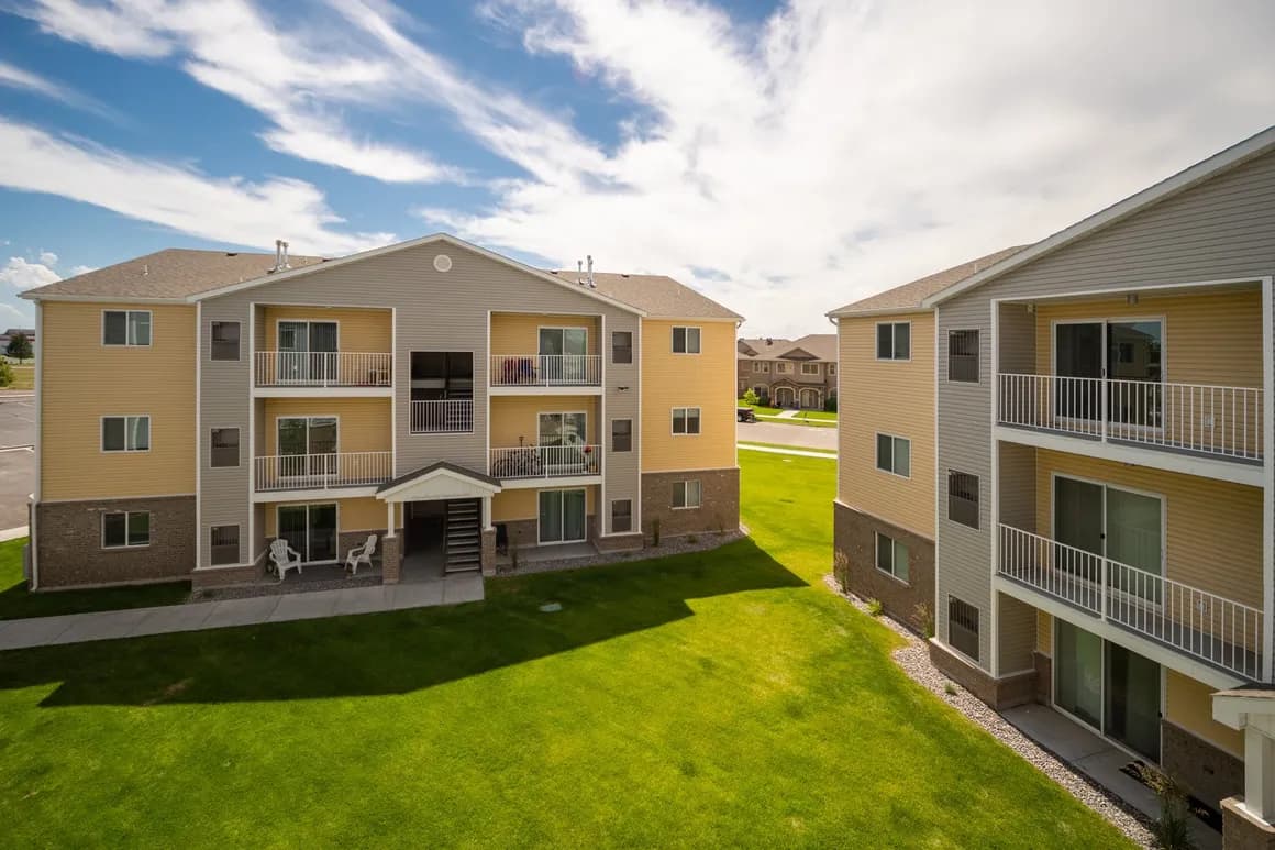 Two modern, three-story apartment buildings at Paddington Place Rexburg with beige siding and white balconies overlook a well-kept green lawn under a partly cloudy sky. Several balconies have outdoor furniture visible.