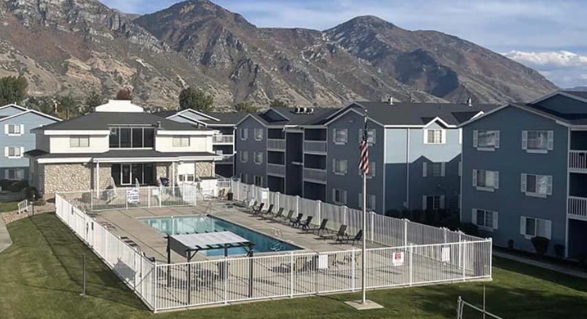 An outdoor swimming pool at Carriage Cove Provo is surrounded by a white fence, nestled between light blue apartment buildings, with mountains in the background under a partly cloudy sky.