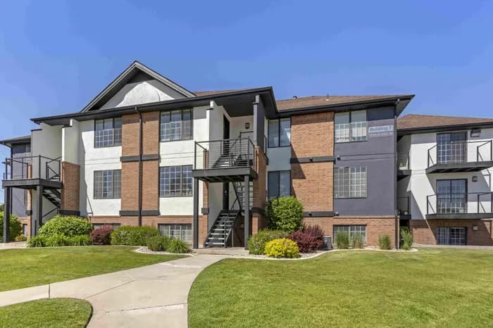 The Branbury Provo offers a two-story apartment building with brown and beige siding, outdoor staircases, and balconies. Several cars are parked in the lot under a clear blue sky.