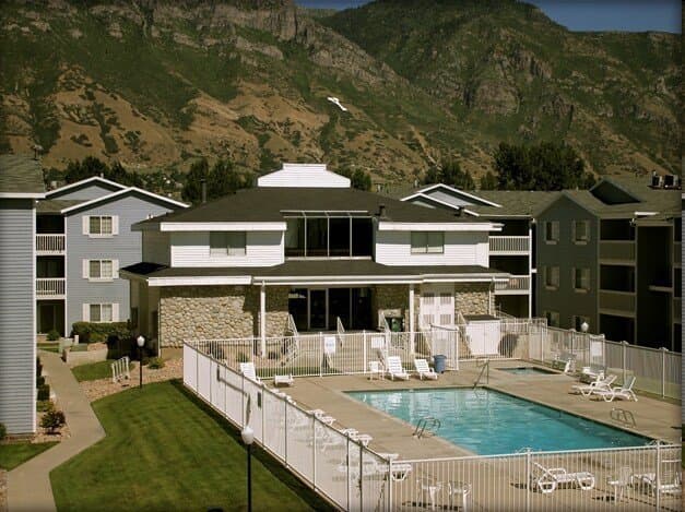 An outdoor swimming pool at Carriage Cove Provo is surrounded by a white fence, nestled between light blue apartment buildings, with mountains in the background under a partly cloudy sky.