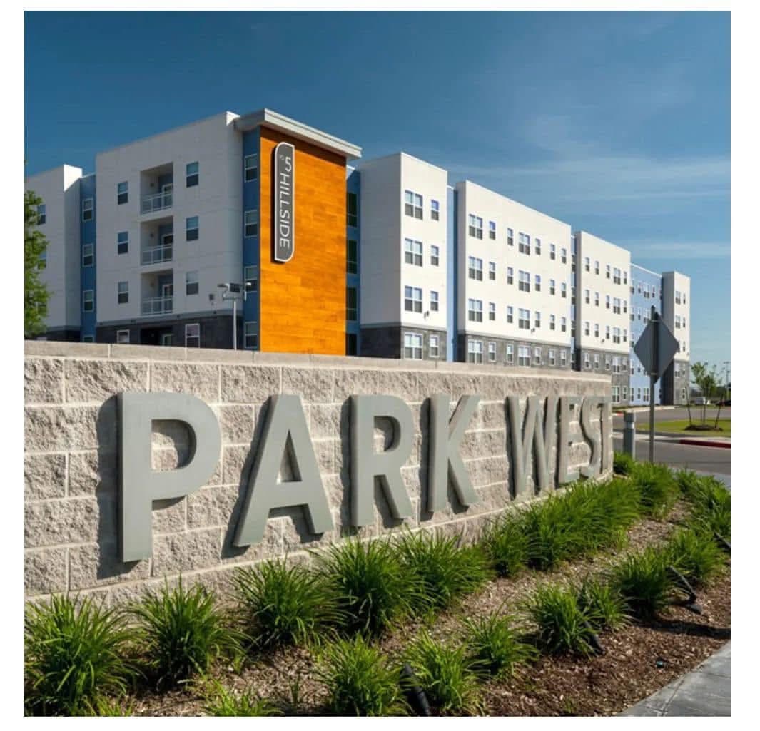 A modern apartment complex with a large outdoor swimming pool, featuring several white lounge chairs partially submerged in the water and rows of poolside seating. The buildings have white, blue, and dark gray exteriors.