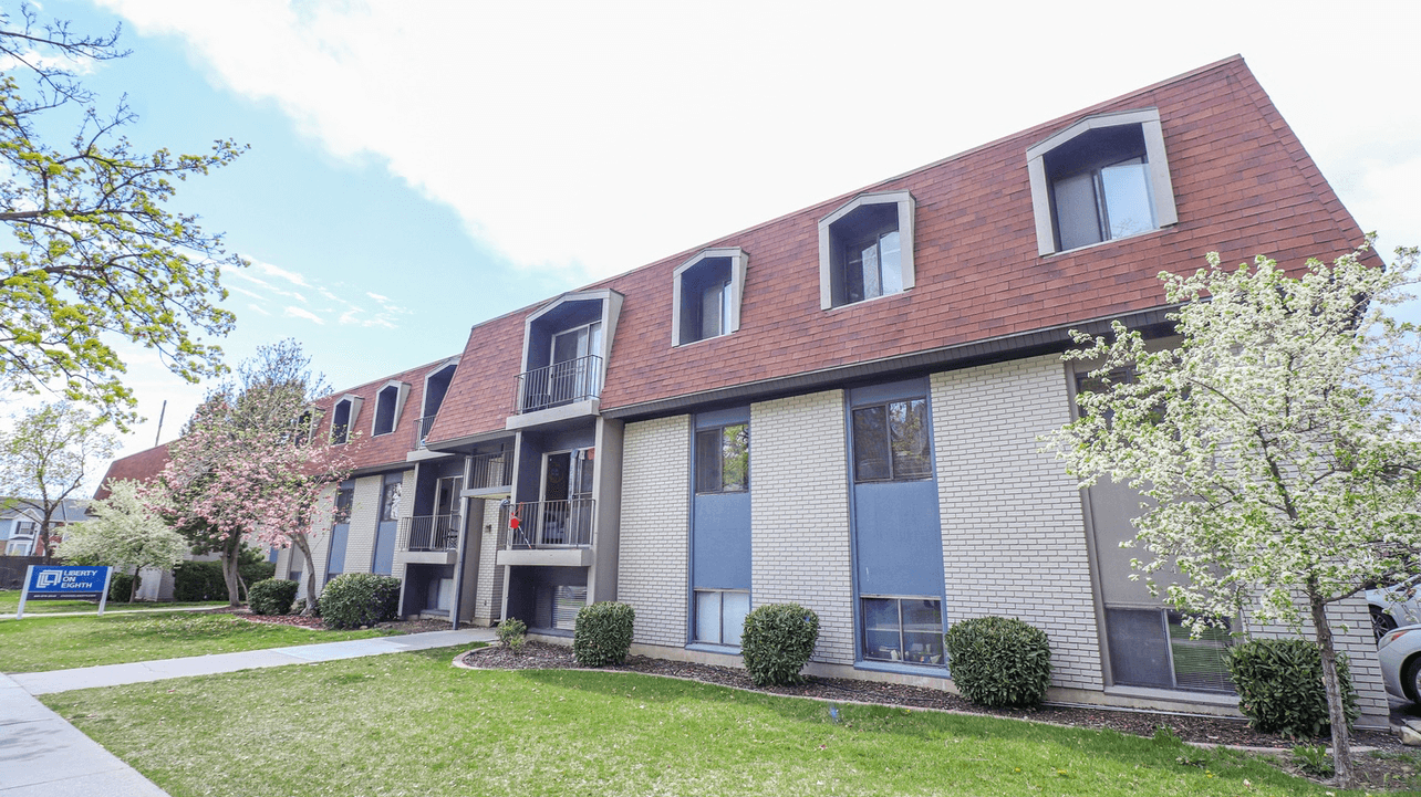 Liberty on Eighth Provo features a three-story apartment building with a brown mansard roof, white and blue-gray exterior walls, several windows, shrubs, a small tree, and a sidewalk in front.