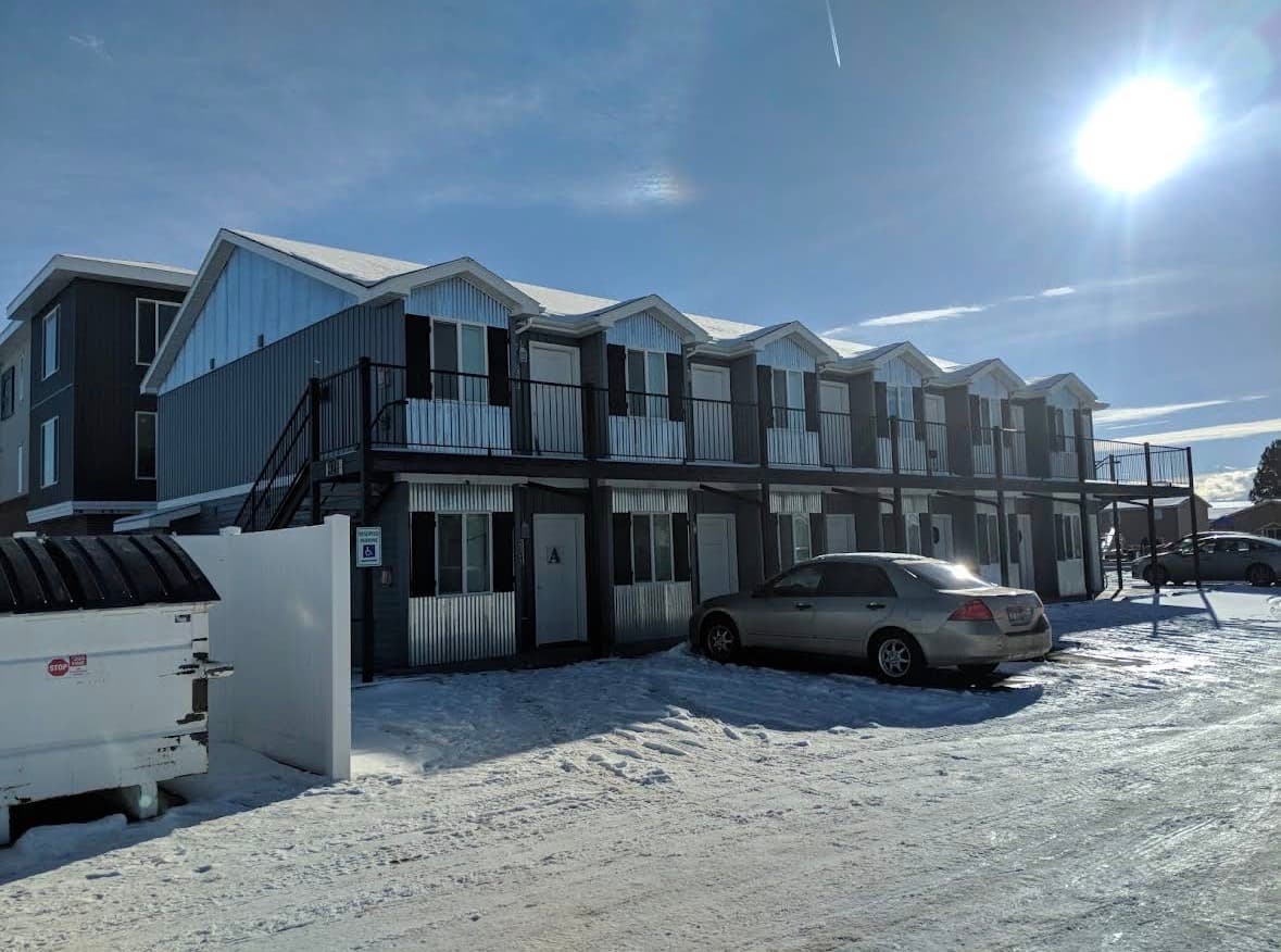 A two-story row of modern, small apartments at Teton Flats Rexburg features white doors, large windows, and a continuous black metal railing along the upper outdoor walkway under a clear sky.