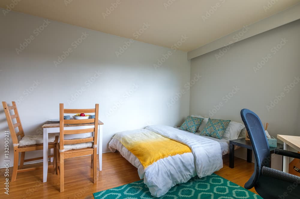 A tidy FM Esatwood court bedroom with a white bed, yellow blanket, two patterned pillows, a small dining table with two chairs, a blue office chair, and a geometric green rug on a wooden floor.