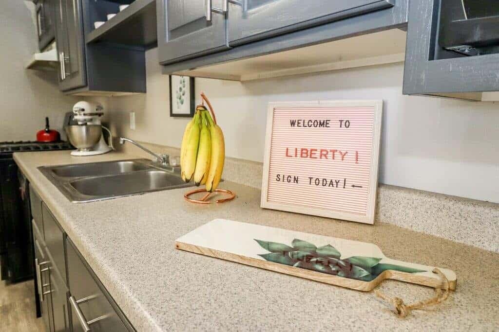 A kitchen countertop at Liberty on Eighth Provo features a bunch of bananas, a cutting board with greenery, and a sign reading WELCOME TO LIBERTY! SIGN TODAY!. A sink and mixer can be seen in the background.