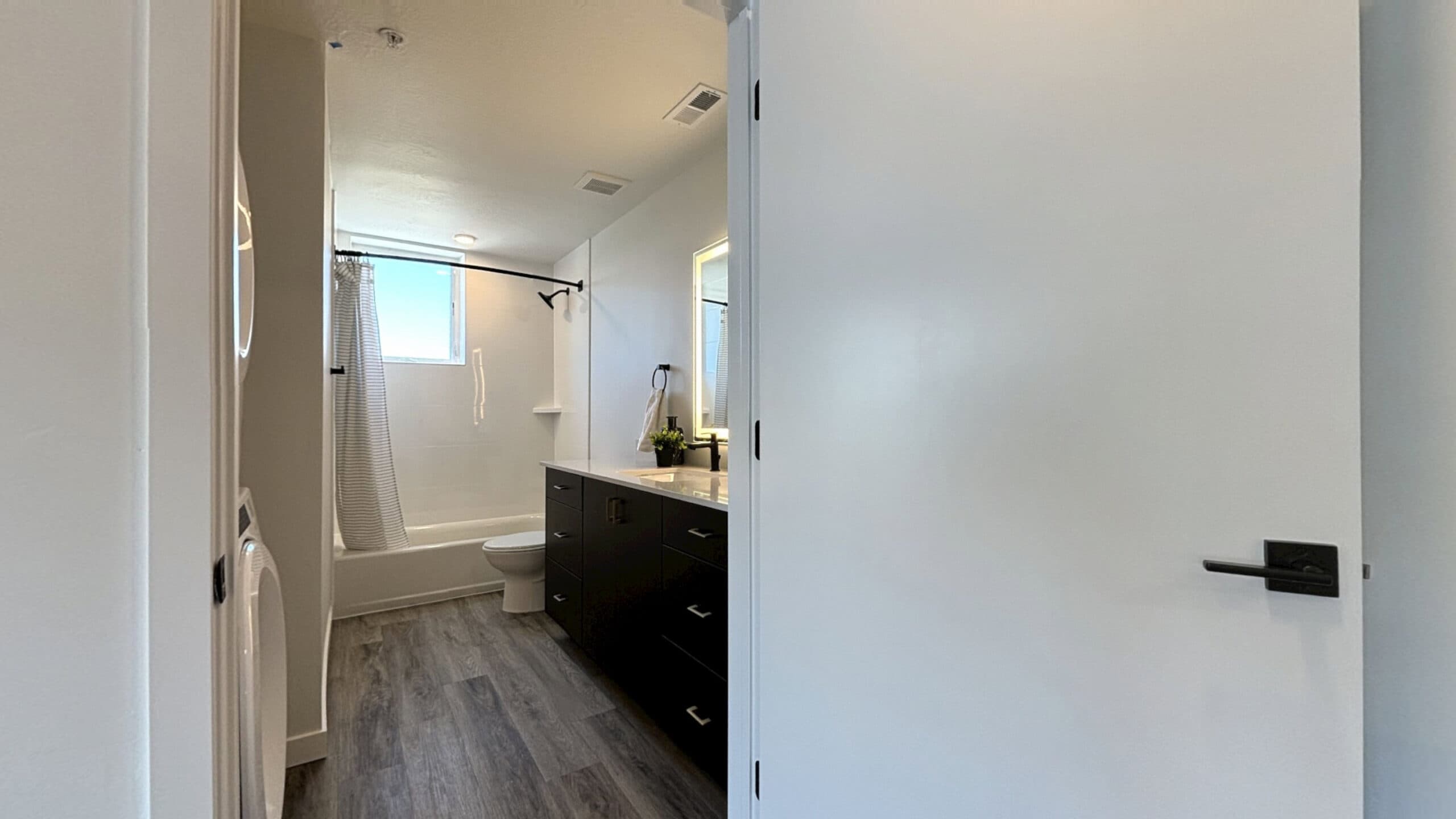 A modern bathroom at 343 Apartments Salt Lake City features wood-look flooring, a white bathtub with shower curtain, toilet, wide vanity with large mirror, and a partially open white door. Natural light streams in from the window.