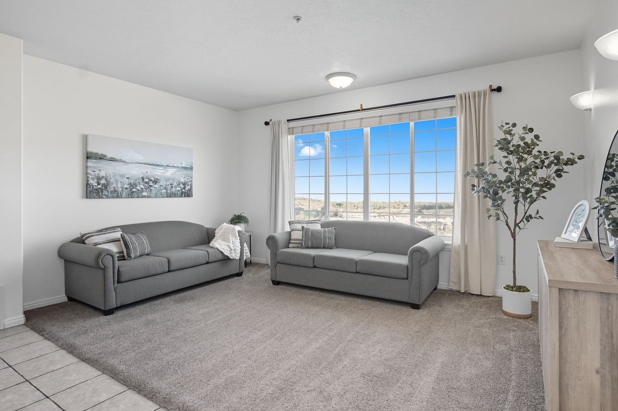 A bright living room at Ventana Student Housing Orem features two gray sofas, a large window with curtains, a potted plant, a landscape painting, and a wooden TV stand on carpeted flooring.