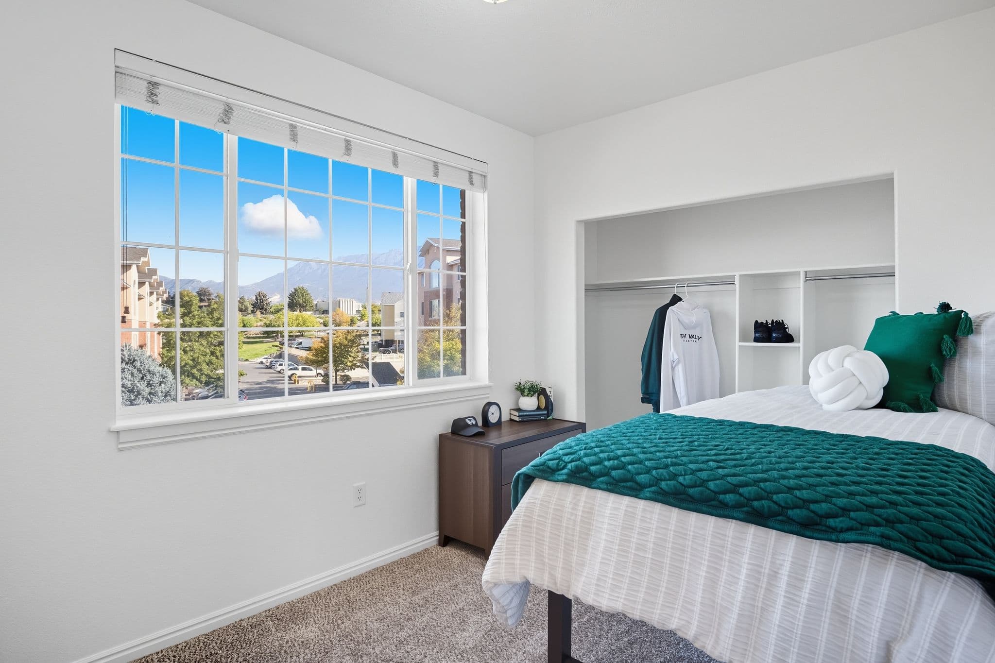 A bright bedroom at Ventana Student Housing Orem features a made bed with green and white bedding, open closet with hanging clothes, small desk, and a large window offering sunny views of apartments and mountains outside.