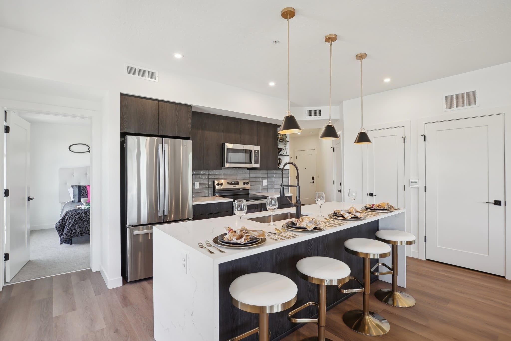 Modern kitchen with dark cabinets, stainless steel appliances, and a white Liberty Duet island with four stools. The island is set for dining with plates and glasses. Pendant lights hang above, and a bedroom is visible through an open door.