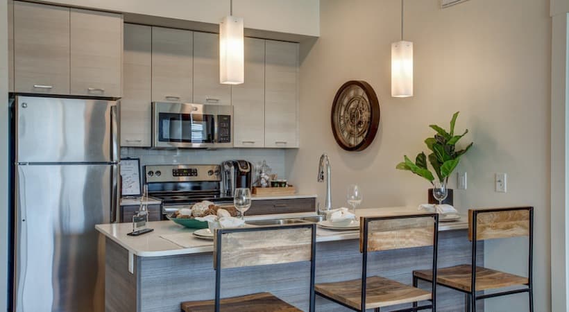 Modern kitchen at Arboretum at Barber Station Boise with light wood cabinets, stainless steel appliances, a kitchen island with three bar stools, pendant lights, potted plant, and decorative wall clock.