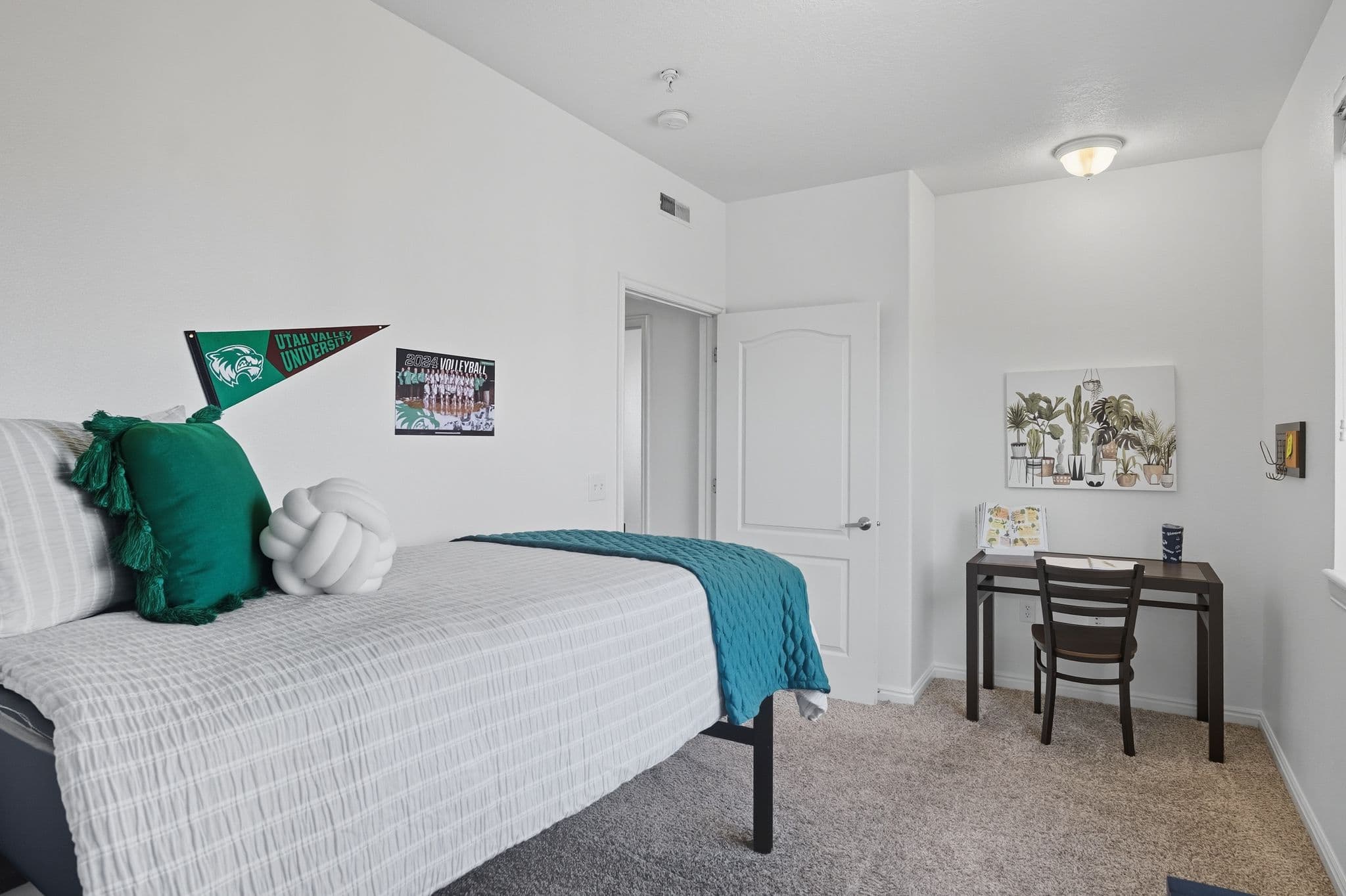 A tidy dorm room at Ventana Student Housing Orem features a neatly made bed with teal accents, a desk with a chair, plant-themed wall art, and university decor on white walls. Natural light brightens the carpeted space.