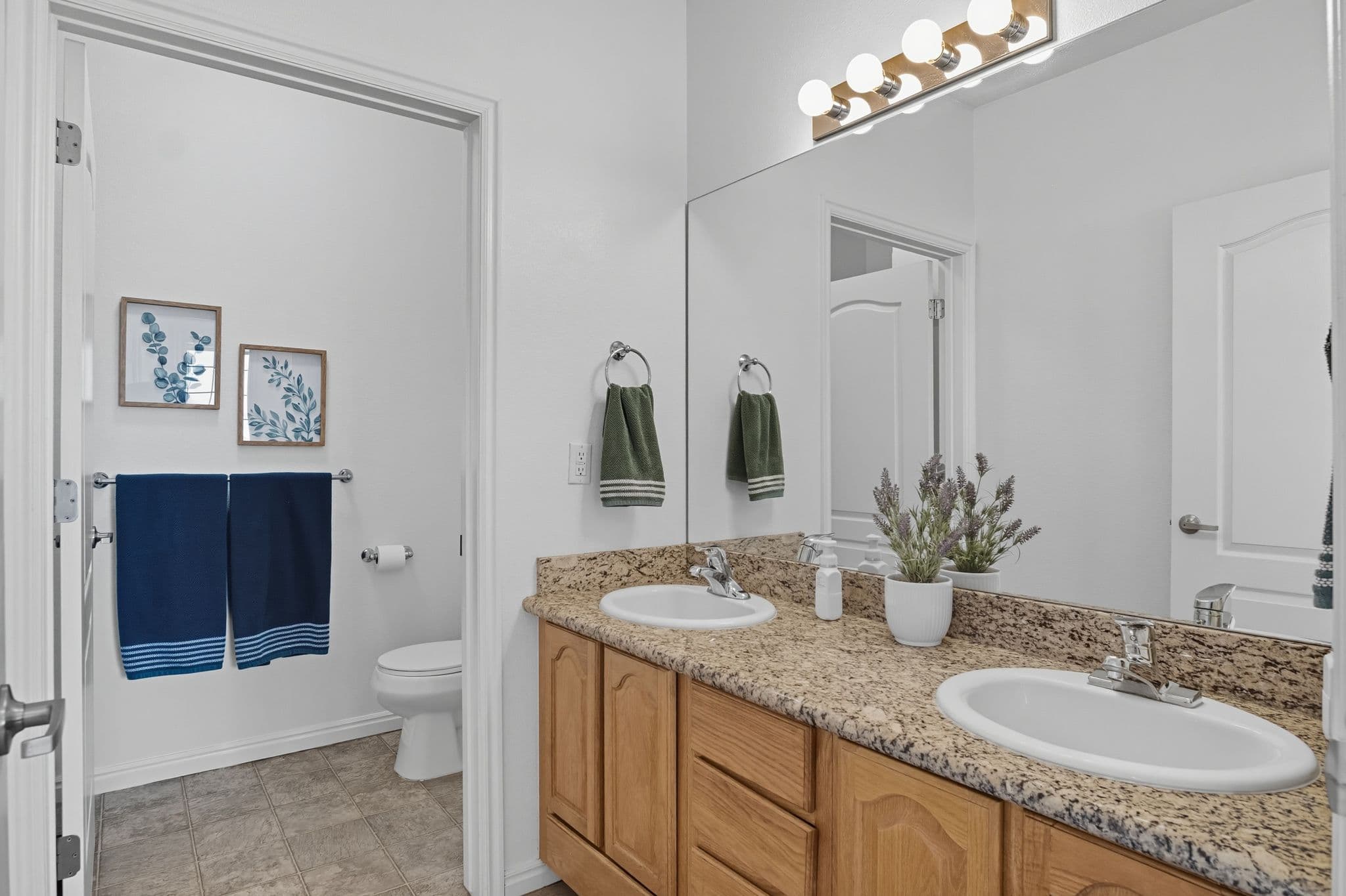 Modern bathroom at Ventana Student Housing Orem with double sinks, granite countertop, wooden cabinets, large mirror, and potted plant. Two blue towels hang near the toilet beneath framed leaf art on the white wall.