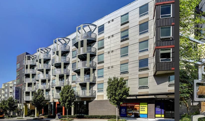 A modern multi-story apartment building in the U District, featuring unique round balconies, large windows, and street-level shops. Several trees line the sidewalk beneath a clear blue sky—welcome to TWELVE.