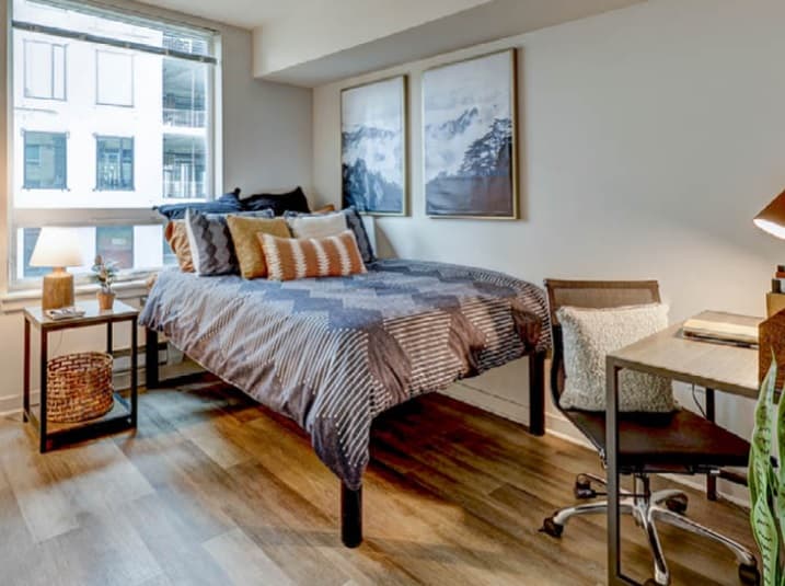 A modern TWELVE bedroom in U District with a neatly made bed, decorative pillows, a desk and chair, small bedside table with lamp, and two framed art pieces on the wall. A large window fills the space with natural light.