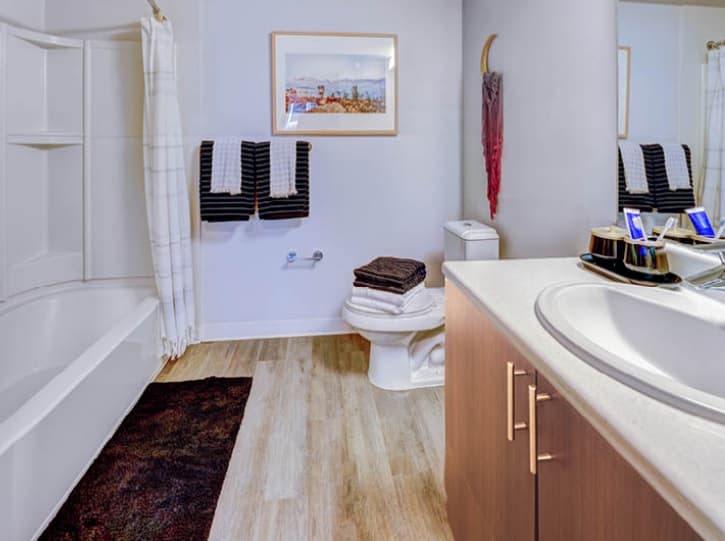 Modern bathroom at TWELVE apartments in Seattle’s U District with a bathtub and shower curtain on the left, toilet with towels stacked on top, vanity with sink and toiletries, wall art above towel racks, and stylish white and wood decor.