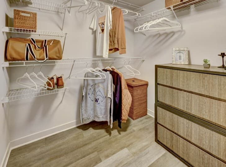 A walk-in closet at TWELVE apartments in the U District features wire shelves with hanging clothes, empty hangers, shoes, a duffel bag, baskets, a framed photo, and a modern wooden dresser on a wood-look floor.