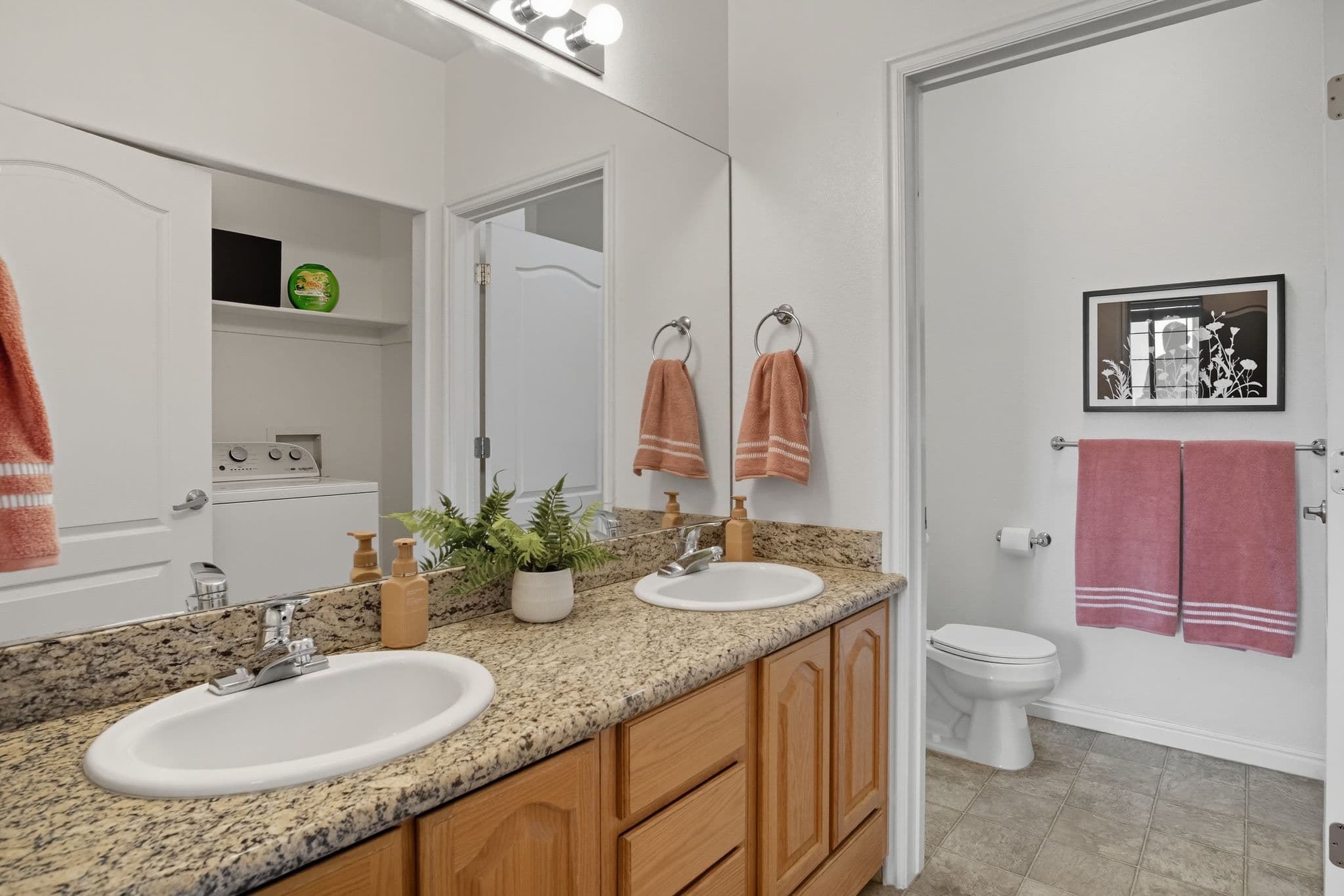 A bathroom at Ventana Student Housing Orem features a granite double sink vanity, wooden cabinets, and a mirror. Pink towels hang on racks, while a potted plant sits on the counter. Doors reveal a toilet with wall art and a laundry area.