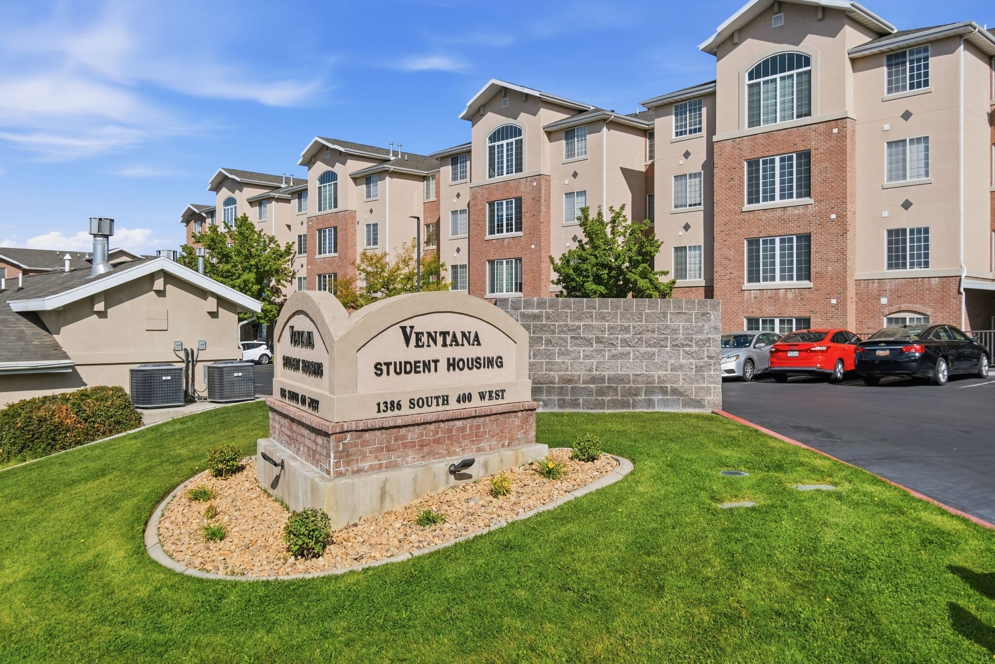 A sign reading “Ventana Student Housing Orem, 1386 South 40th West” stands on a landscaped lawn in front of a multi-story apartment building with red brick and beige siding. Several cars are parked nearby.