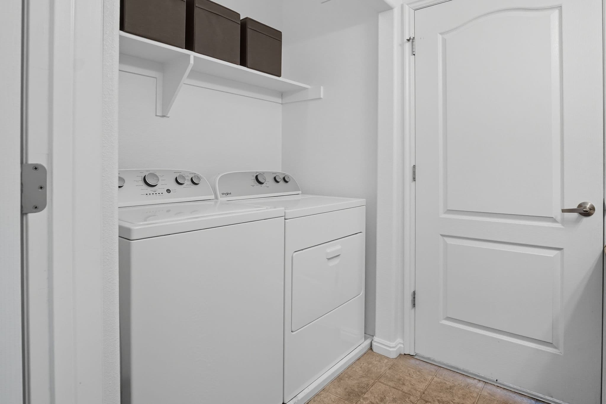 A laundry room at Ventana Student Housing Orem features a white washing machine and dryer side by side, a white door to the right, and a wall shelf above with three dark storage bins. The light brown tiled floor adds warmth to the space.