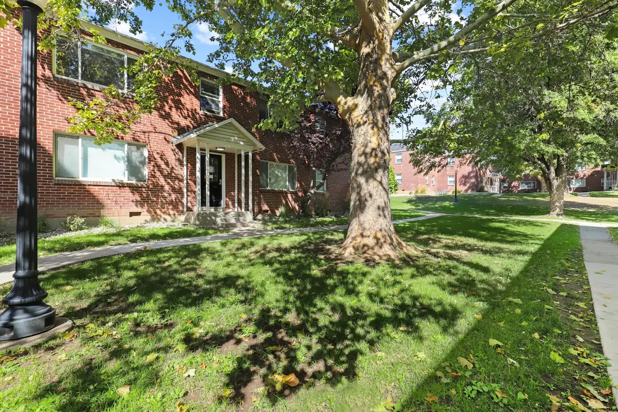 A brick apartment building with large windows at University Gardens Apartments in Salt Lake City is shaded by tall trees on a grassy lawn, with a sidewalk and lamppost visible in the foreground.