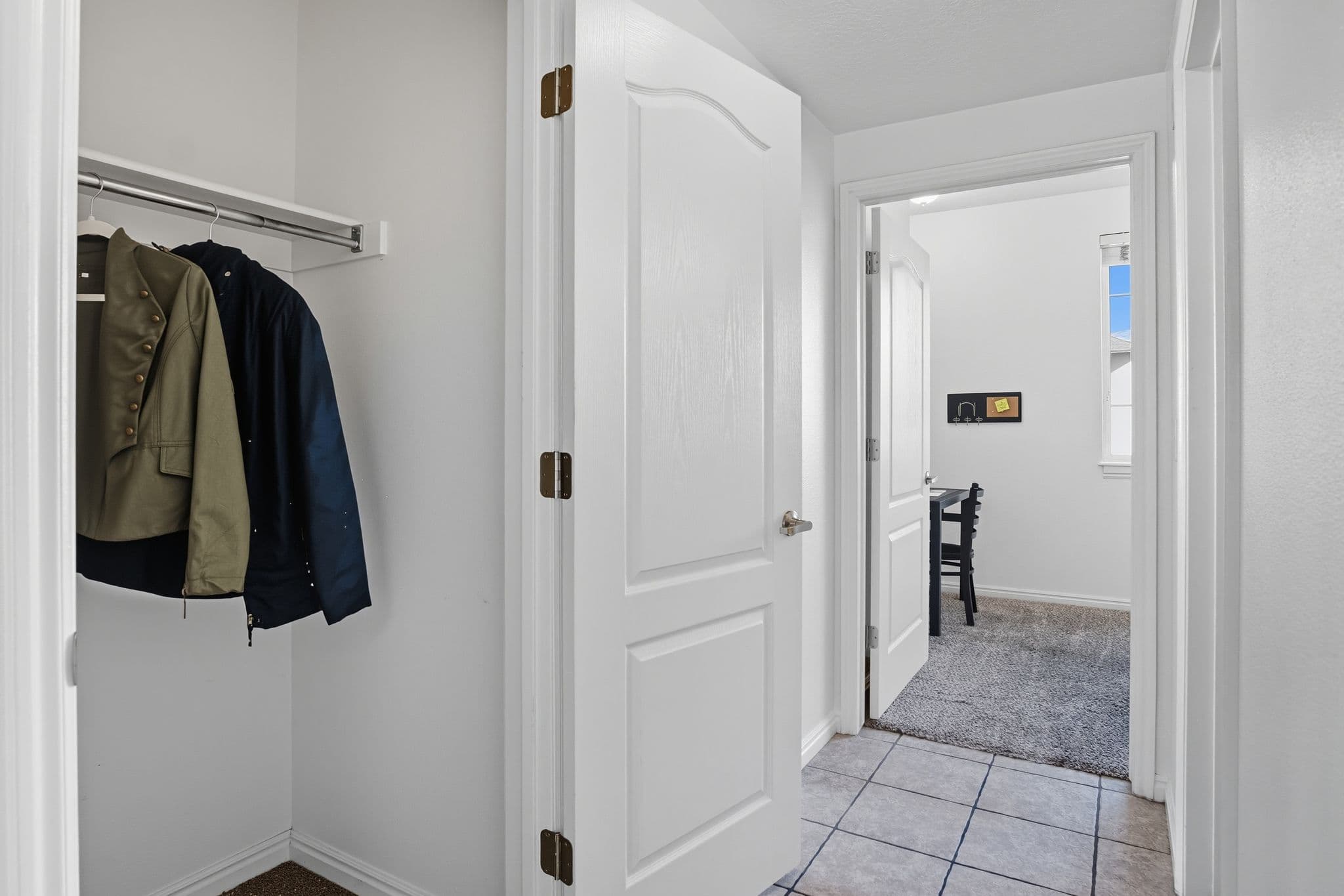 A hallway at Ventana Student Housing Orem features white walls and doors, a tile floor, and an open closet on the left with a green and blue jacket. Through the open door ahead, a small office with a desk and chair is visible.