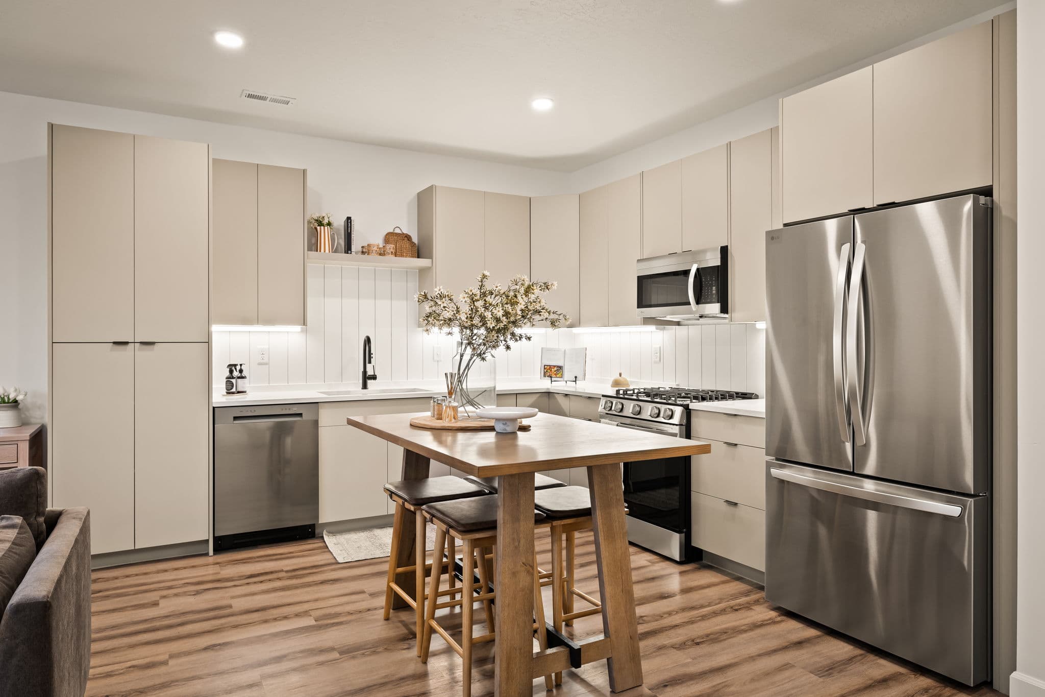 Modern kitchen with beige cabinets, stainless steel appliances, a wooden island with stools, a vase with flowers, and bright lighting. The space features light wood flooring and minimalist decor.