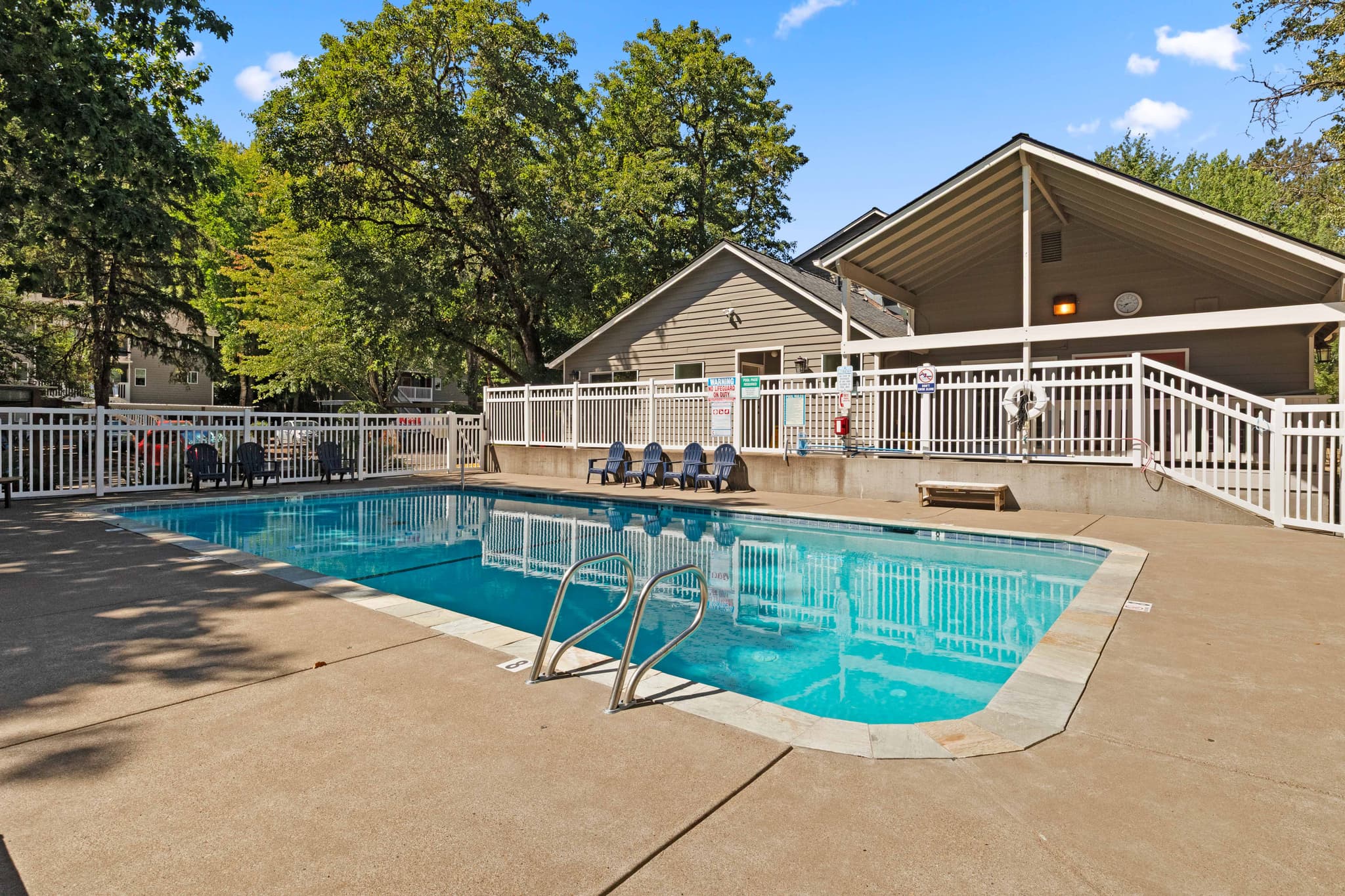 Outdoor swimming pool with metal handrails, surrounded by a concrete deck and white fence at Witham Village. Several lounge chairs and a beige clubhouse building sit in the background amid green trees and blue sky.