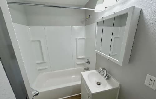 A small, modern bathroom with a white bathtub and shower, a white vanity with a sink, a mirrored medicine cabinet, and light gray walls.