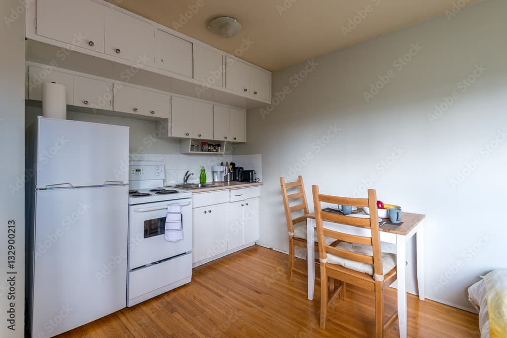 A small kitchen with white cabinets, a stove, refrigerator, and countertop appliances. Next to it is a wooden dining table with two chairs on a hardwood floor, all set against plain white walls in FM Eastwood Court.
