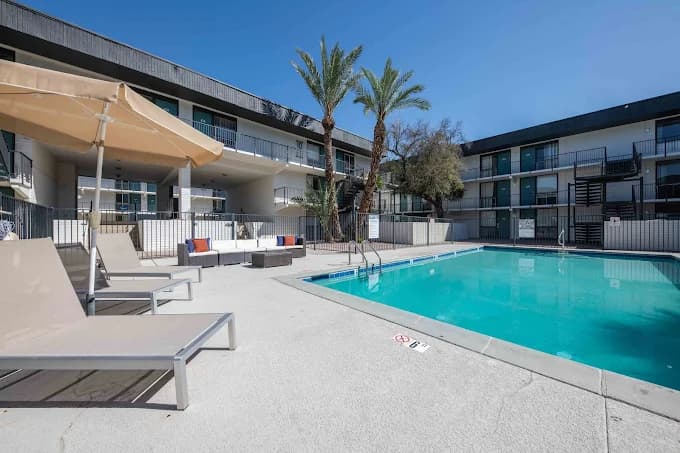 Outdoor swimming pool surrounded by lounge chairs and a large umbrella, with Spectra Midtown Apartments Phoenix rising in the background and palm trees swaying beneath a clear blue sky.