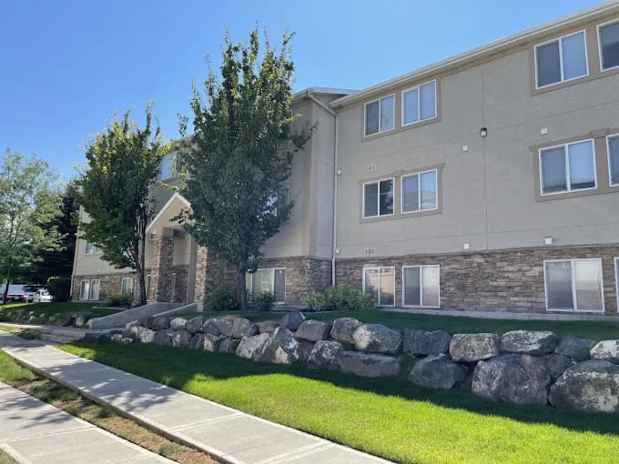 The Ridge Rexburg is a three-story apartment building with beige walls and stone accents, surrounded by green grass, trees, and large rocks along a sidewalk under a clear blue sky.