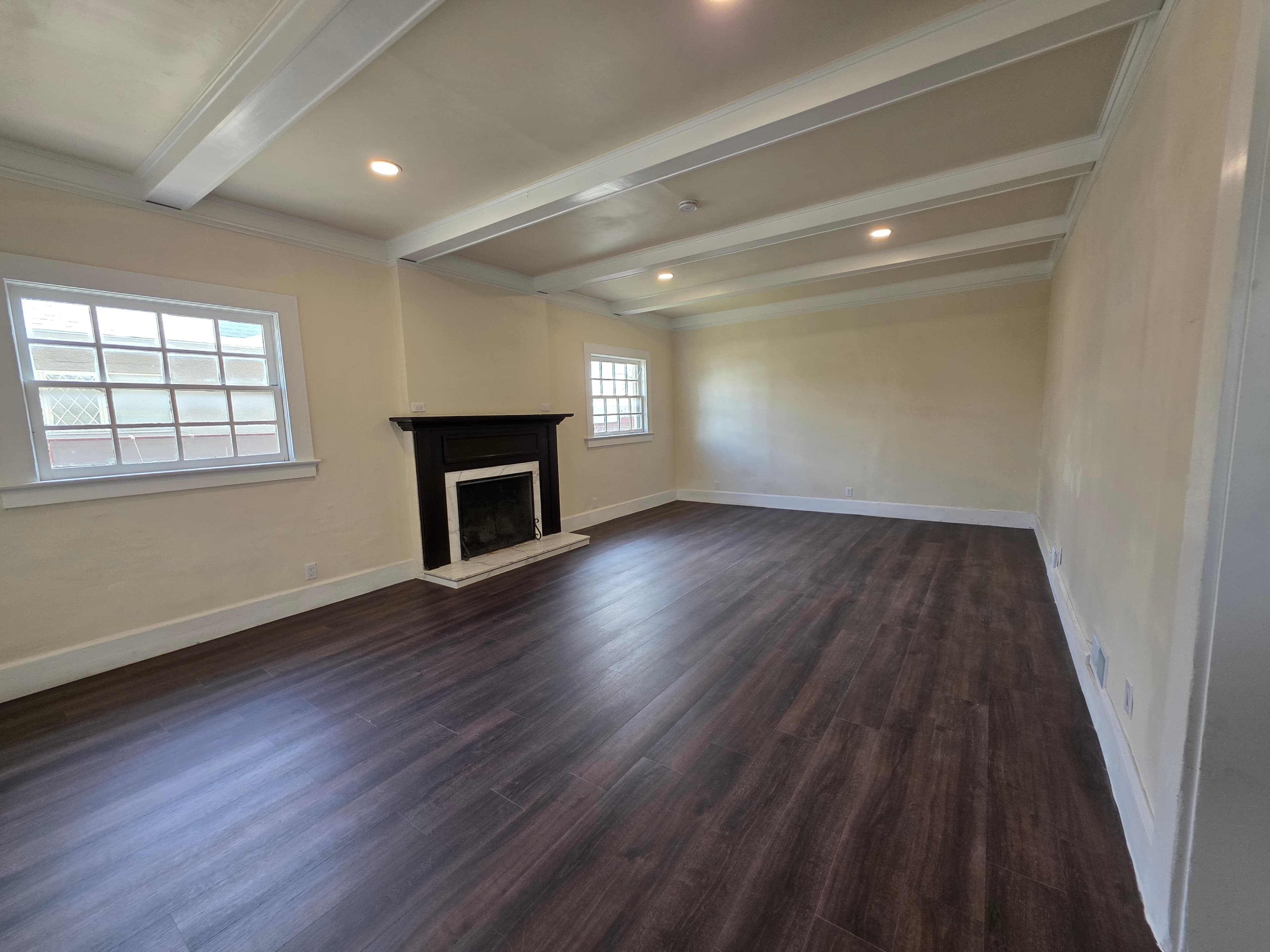 A spacious, empty living room at 833 N 4th Ave features dark wood flooring, cream-colored walls, a white beamed ceiling, two windows, and a black fireplace with a white mantle. Recessed lighting brightens the room.