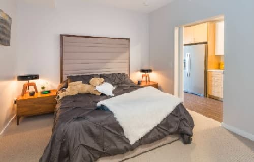 A modern bedroom with a large bed covered in dark bedding, a tan headboard, two bedside tables with lamps, a teddy bear on the bed, and an open doorway leading to a kitchen area.