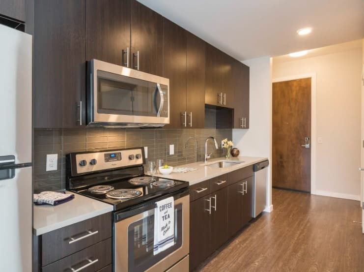 Modern kitchen with dark wood cabinets, stainless steel appliances, a microwave above an electric stove, a sink, and a countertop with a coffee and tea setup. The floor is wood, and the space looks clean and bright.