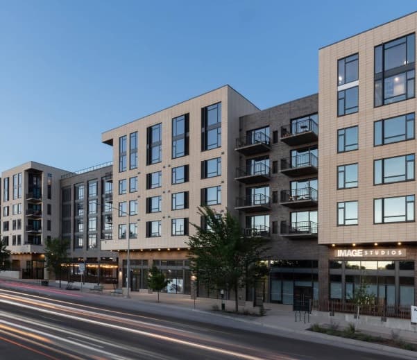 Modern mid-rise apartment building with large windows and balconies, retail shops at street level, and blurred car lights passing by on the road at dusk.