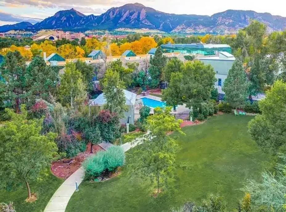 Aerial view of The View on 26th Boulder: a green park with walking paths, trees, and a swimming pool. Buildings are partly visible among the trees, with mountains and autumn foliage in the background under a partly cloudy sky.