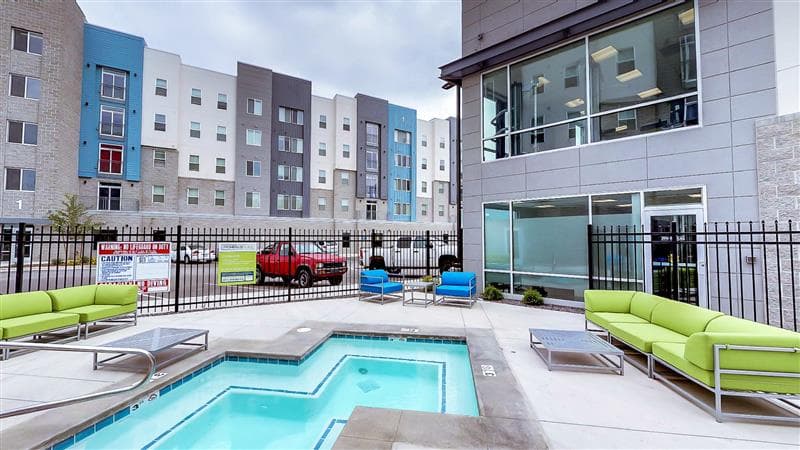 A small outdoor pool with clear water is surrounded by green and blue lounge seating at Promenade Place Orem. Modern apartment buildings and a red vehicle are visible in the background behind a black metal fence.