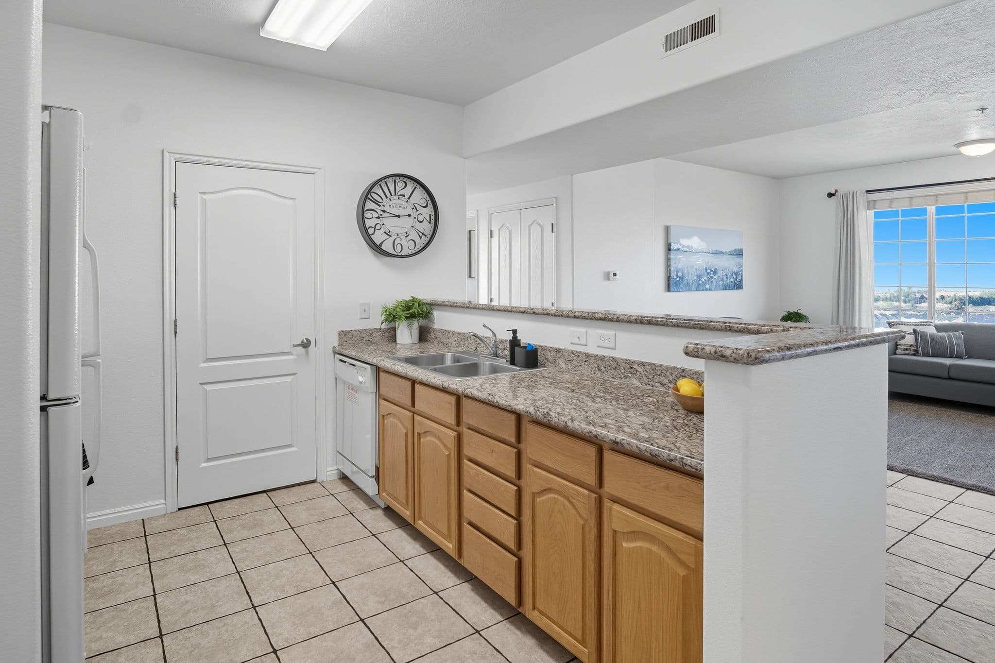 A modern kitchen at Ventana Student Housing Orem features light wood cabinets, granite countertops, and a double sink overlooking a bright living room with a large window. A wall clock, potted plant, and bowl of lemons add decorative touches.