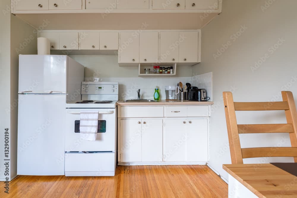 Small kitchen at FM Eastwood Court with white cabinets, a white refrigerator and stove, wooden floor, countertop appliances, and a partially visible wooden chair in the foreground. The space appears tidy and compact.