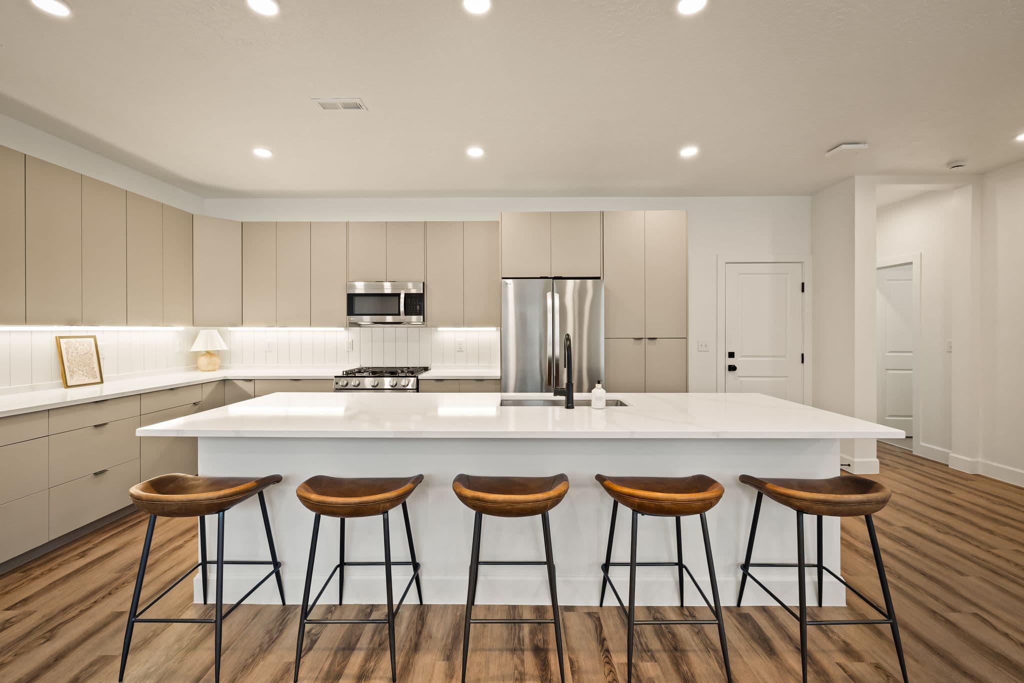 Modern kitchen with beige cabinets, stainless steel appliances, white countertops, and a large island with five brown wooden bar stools. Wooden flooring and recessed ceiling lights create a bright, welcoming space.