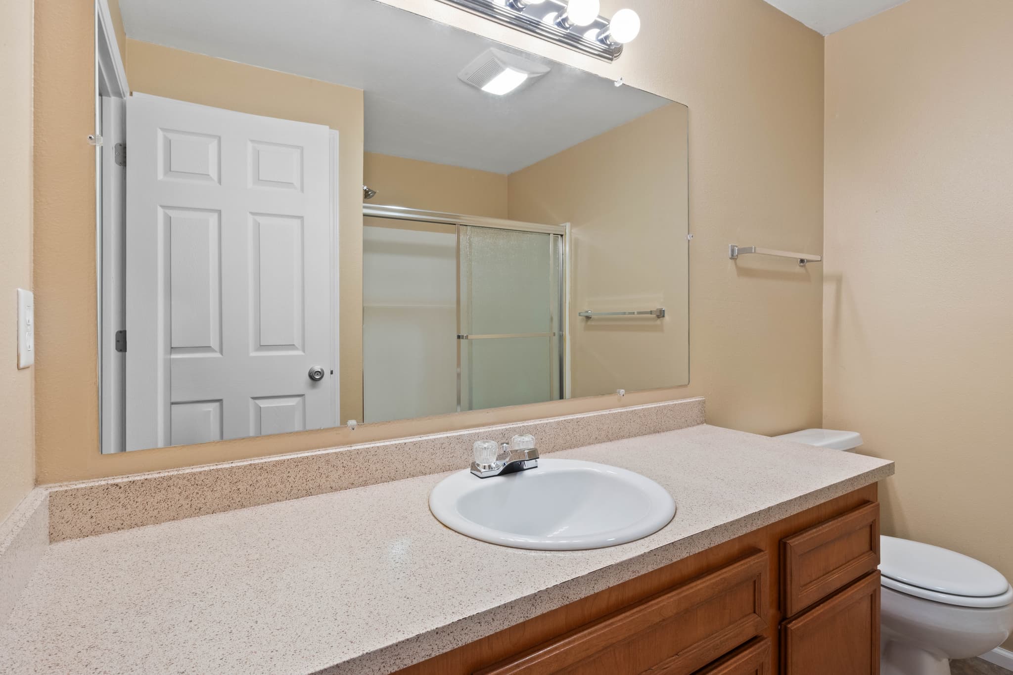 A clean bathroom in Witham Village features a beige countertop, white sink, wooden cabinets, large mirror, white toilet, and a shower with sliding glass doors. The light tan walls and lighting above the mirror create a bright, welcoming space.