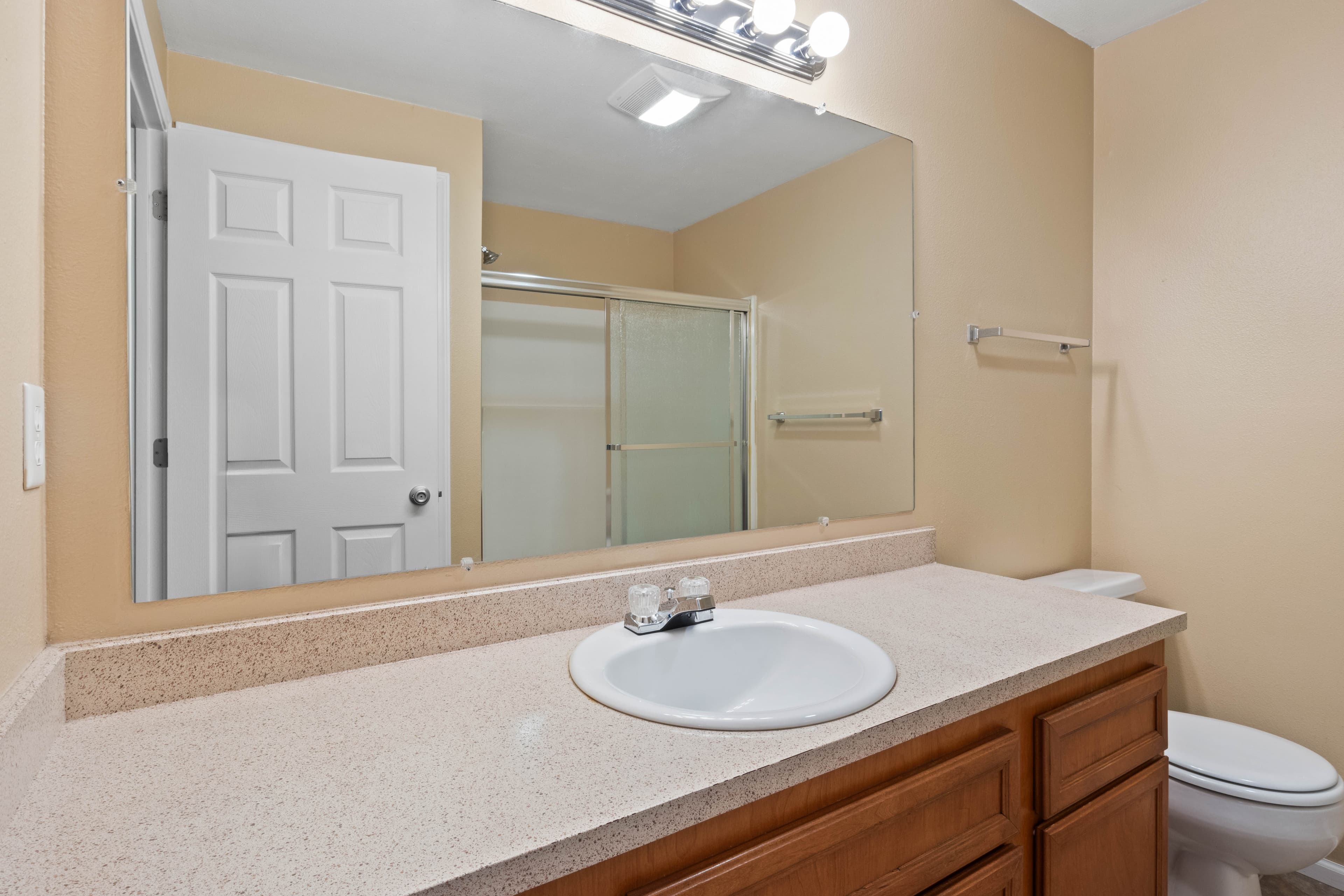 A clean bathroom in Witham Village features a beige countertop, white sink, wooden cabinets, large mirror, white toilet, and a shower with sliding glass doors. The light tan walls and lighting above the mirror create a bright, welcoming space.