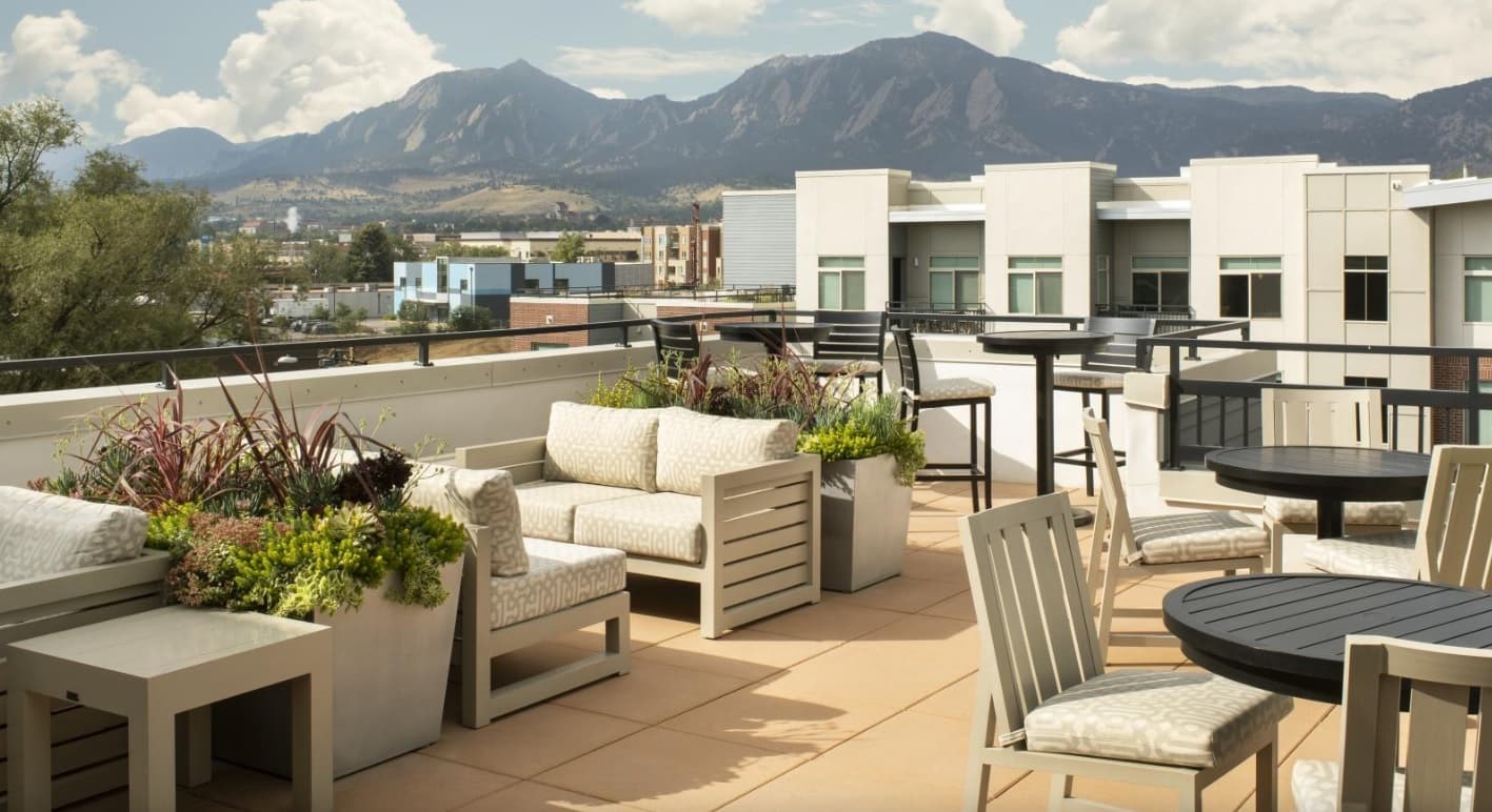 Rooftop patio at 3100 Pearl Boulder features cushioned seating, tables, potted plants, and modern railings overlooking city buildings and distant mountains under a partly cloudy sky.