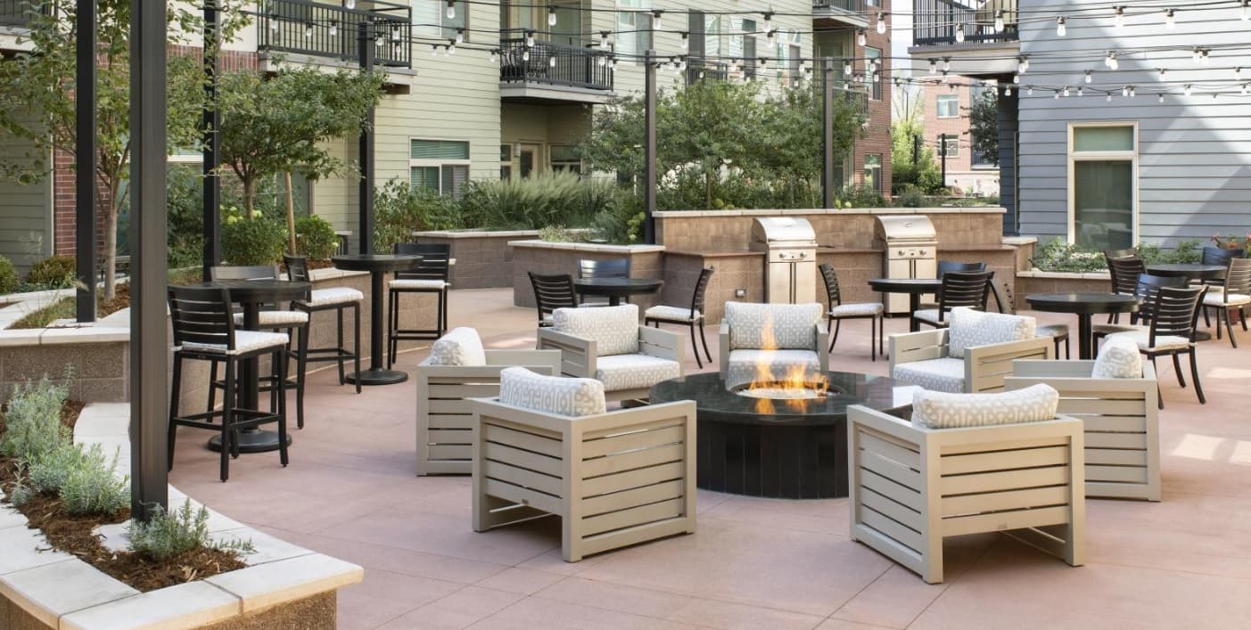 Outdoor patio area at 3100 Pearl Boulder features cushioned chairs around a fire pit, nearby tables and barstools, string lights overhead, built-in grills, and plants along the edges, all surrounded by apartment buildings.