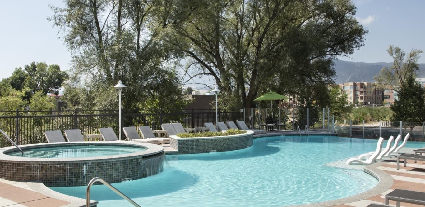 Outdoor pool with curved edges and a connected circular hot tub at 3100 Pearl Boulder, surrounded by lounge chairs, trees, and an umbrella-covered table. City buildings provide a scenic backdrop for relaxing moments.