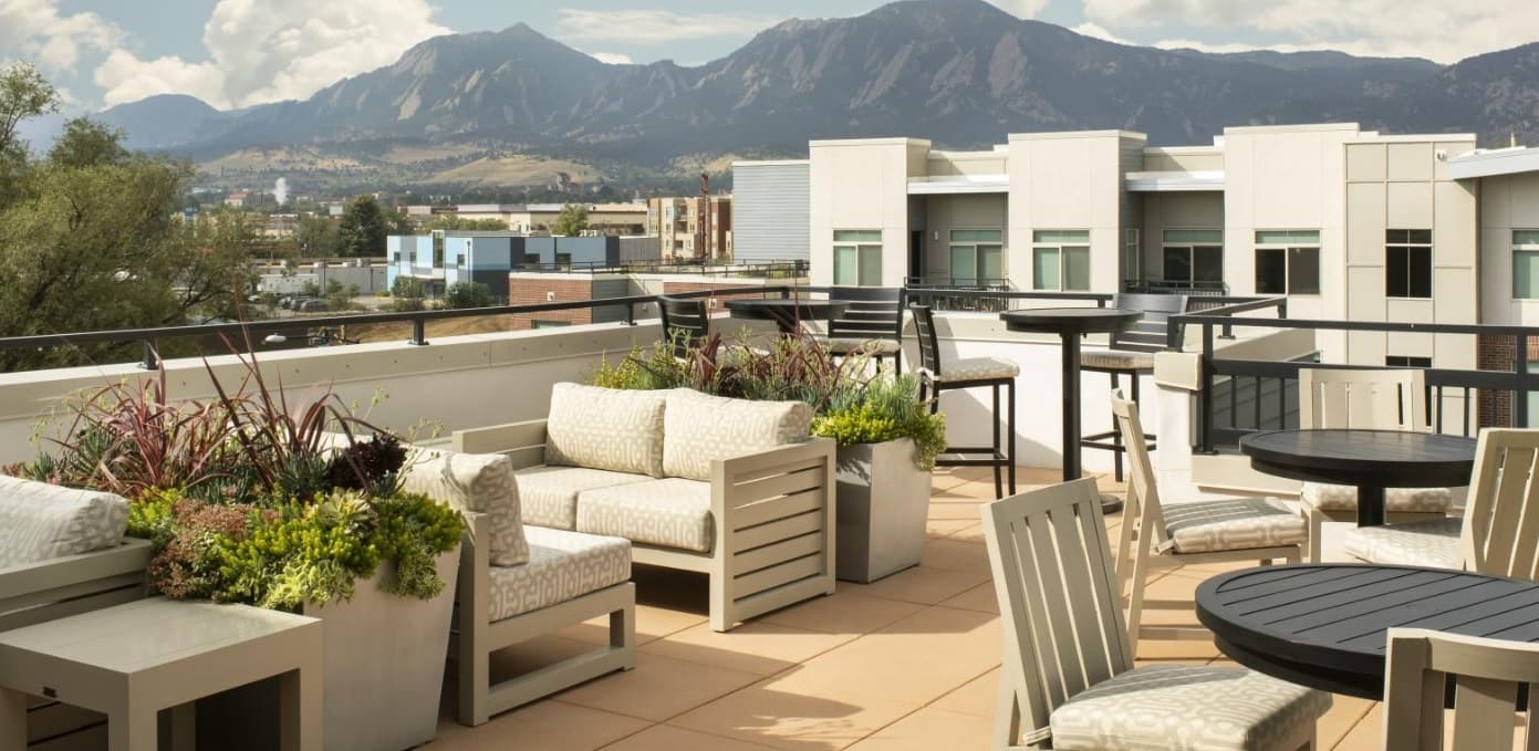 A modern rooftop patio at 3100 Pearl Boulder features cushioned seating, potted plants, and round tables, overlooking a cityscape and distant mountains under a partly cloudy sky.
