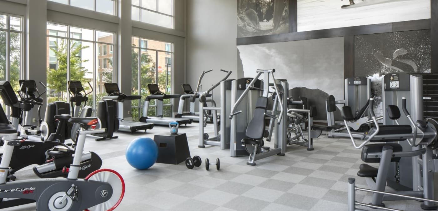 A modern gym at 3100 Pearl Boulder features large windows, various exercise machines, stationary bikes, treadmills, free weights, a blue exercise ball, and bright natural lighting. Monochrome fitness-themed artwork decorates the walls.