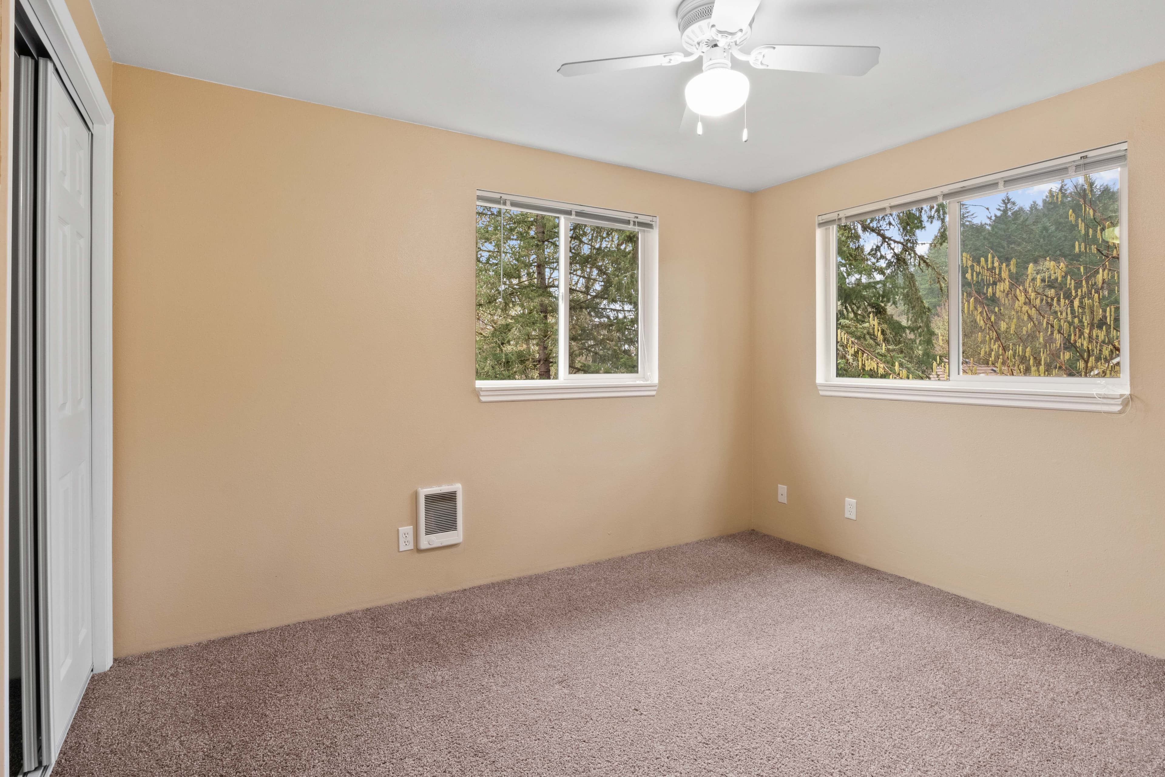 An empty bedroom in Witham Village features beige walls, light brown carpet, two windows with views of trees, a white ceiling fan with light, and a sliding closet door.